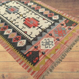 May include: A colorful rug with a geometric pattern in shades of red, black, white, and blue. The rug has a fringe border and is made of wool.