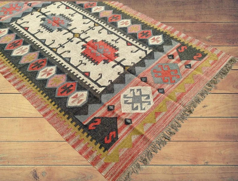 May include: A colorful rug with a geometric pattern in shades of red, black, white, and blue. The rug has a fringe border and is made of wool.