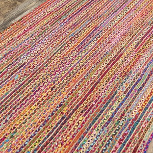 May include: A rectangular braided rug with a vibrant, multicolored striped pattern. The rug features a mix of red, pink, orange, yellow, green, blue, and purple hues. The rug is made of woven material and is placed on a wooden floor.