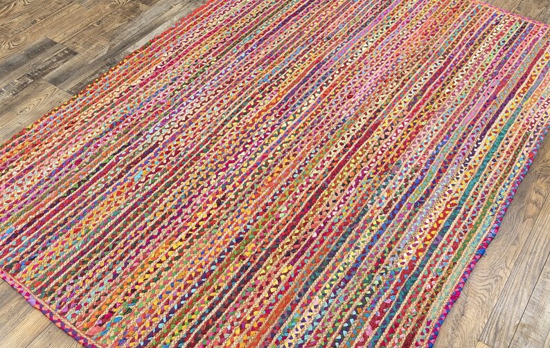 May include: A rectangular braided rug with a vibrant, multicolored striped pattern. The rug features a mix of red, pink, orange, yellow, green, blue, and purple hues. The rug is made of woven material and is placed on a wooden floor.