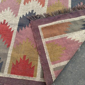 May include: A colorful geometric patterned rug with a fringe border. The rug is made of woven fabric and features shades of red, brown, gray, and white.