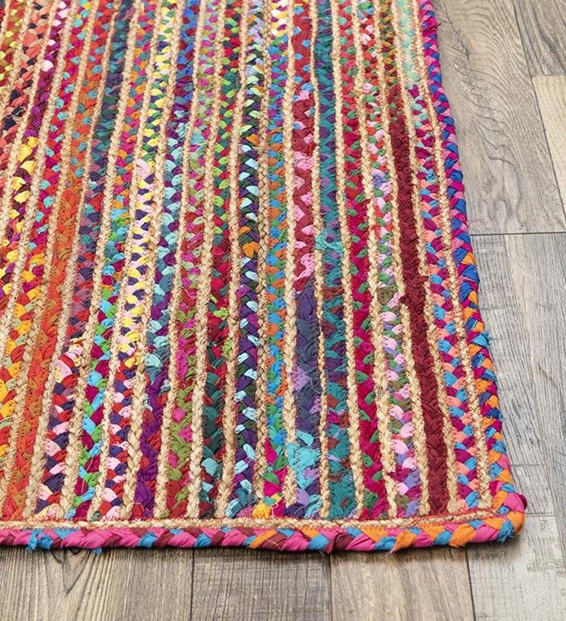 May include: A colorful braided rug with vertical stripes in shades of red, pink, orange, yellow, green, blue, and purple. The rug has a braided border in pink and orange. The rug is made of a natural fiber material.