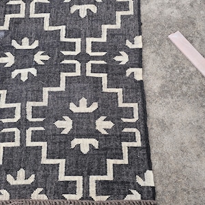 May include: A dark gray and white patterned rug with a geometric design. The rug features a repeating pattern of stepped lines and arrow-like shapes. A light-colored wooden piece is next to the rug.