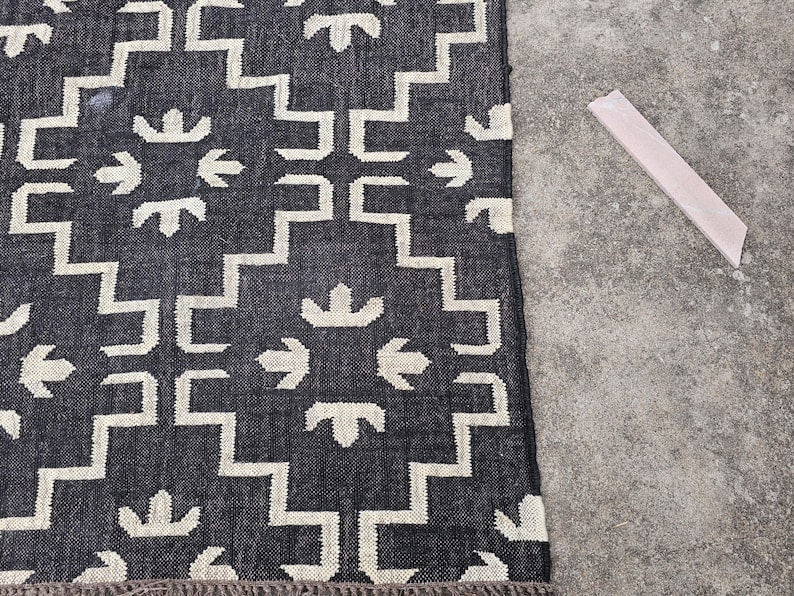May include: A dark gray and white patterned rug with a geometric design. The rug features a repeating pattern of stepped lines and arrow-like shapes. A light-colored wooden piece is next to the rug.