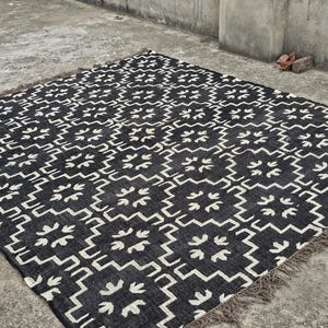 May include: A black and white patterned rug with a geometric design. The rug features a repeating pattern of stylized floral shapes and zigzags. The rug has a fringed edge and is placed on a concrete surface.