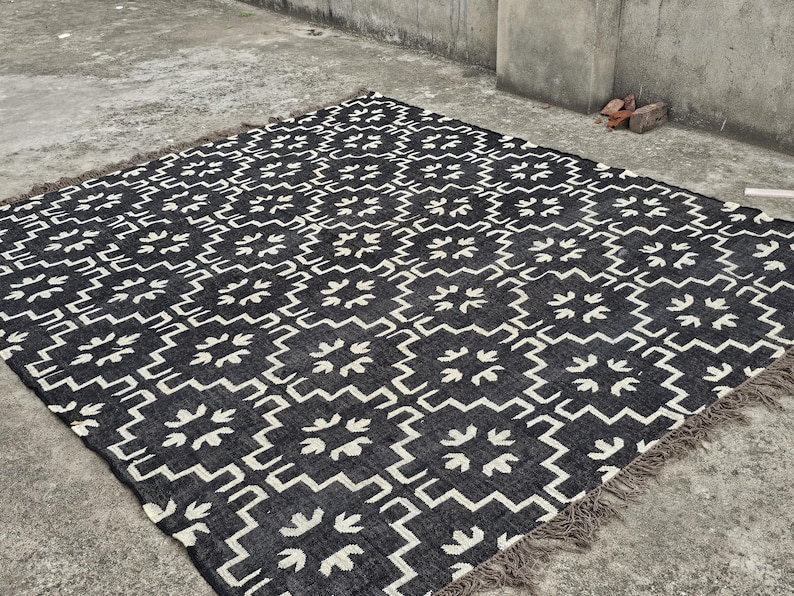 May include: A black and white patterned rug with a geometric design. The rug features a repeating pattern of stylized floral shapes and zigzags. The rug has a fringed edge and is placed on a concrete surface.