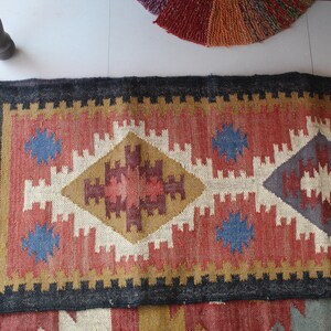 May include: A colorful rug with a geometric pattern in shades of red, blue, white, and brown. The rug has a black border and fringe on the ends.