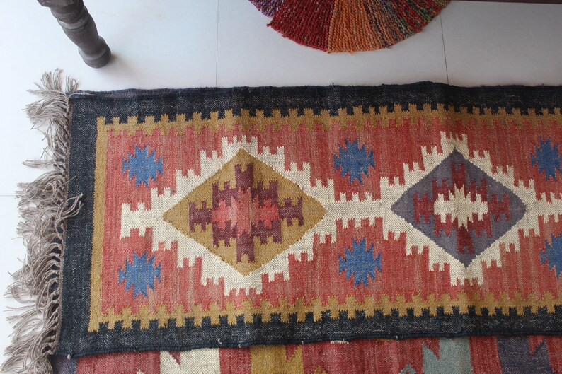 May include: A colorful rug with a geometric pattern in shades of red, blue, white, and brown. The rug has a black border and fringe on the ends.