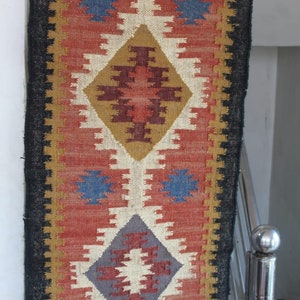 May include: A long, narrow rug with a geometric pattern in shades of red, brown, white, blue, and gray. The rug has a black border and a fringe on the top and bottom.