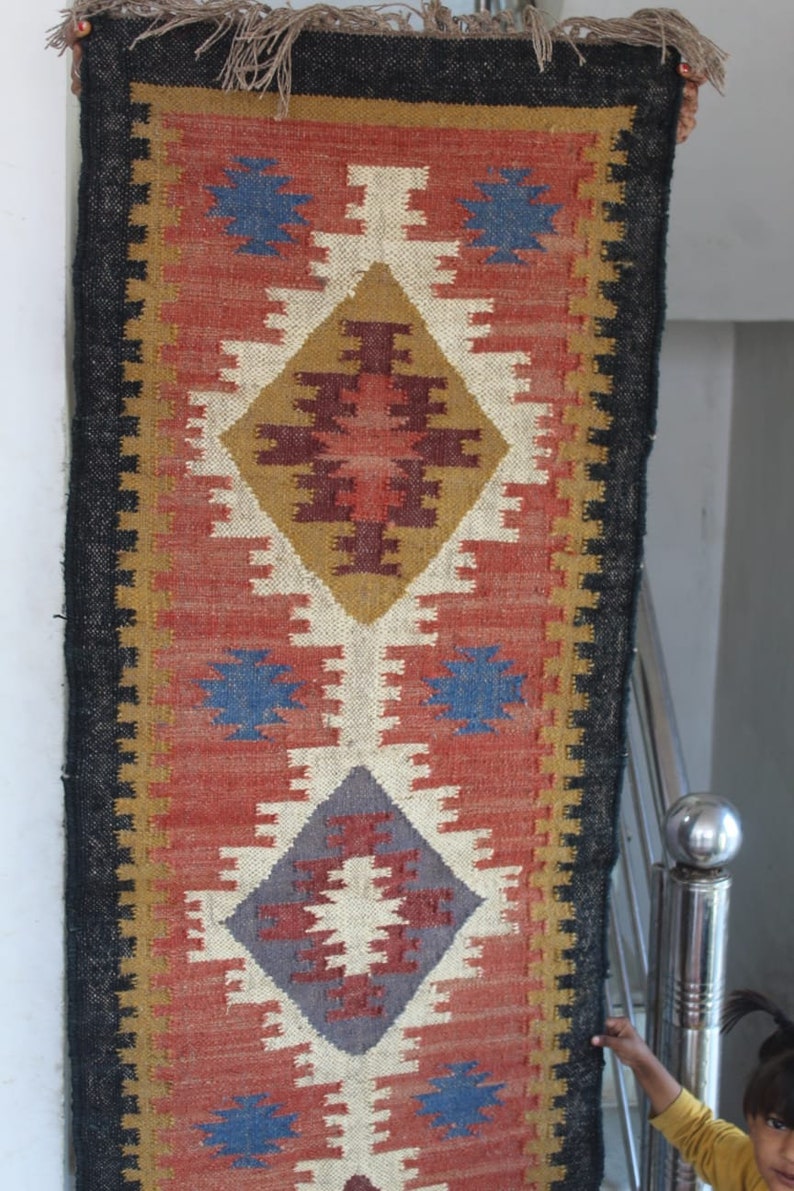 May include: A long, narrow rug with a geometric pattern in shades of red, brown, white, blue, and gray. The rug has a black border and a fringe on the top and bottom.