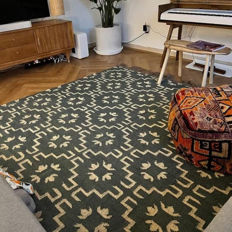 May include: A dark green and cream patterned rug with a geometric design. The rug is laid out on a hardwood floor in a living room setting. A colorful pouf sits on the rug.