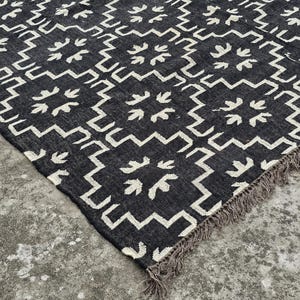 May include: A black and white patterned rug with a geometric design. The rug features a repeating pattern of white shapes on a black background. The edges have a fringe detail.