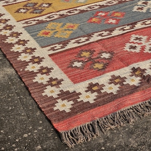 Può includere: Tappeto fantasia con un design geometrico nei toni del rosso, blu e giallo. Il tappeto ha un bordo con motivi floreali ripetuti e un bordo con frange. Realizzato in tessuto, ideale per soggiorno o camera da letto.