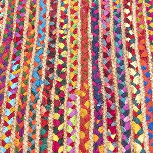 May include: Close-up of a colorful braided rug with vertical stripes. The rug features a mix of vibrant colors including red, blue, yellow, and green. The braided sections are separated by natural-colored jute. This rug is suitable for home decor.