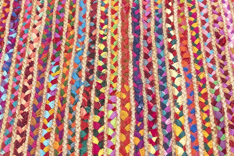 May include: Close-up of a colorful braided rug with vertical stripes. The rug features a mix of vibrant colors including red, blue, yellow, and green. The braided sections are separated by natural-colored jute. This rug is suitable for home decor.