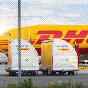 May include: A yellow DHL cargo plane with red lettering is parked on the tarmac. In front of the plane are several white and yellow cargo containers with the DHL logo. A chain-link fence is in the foreground.
