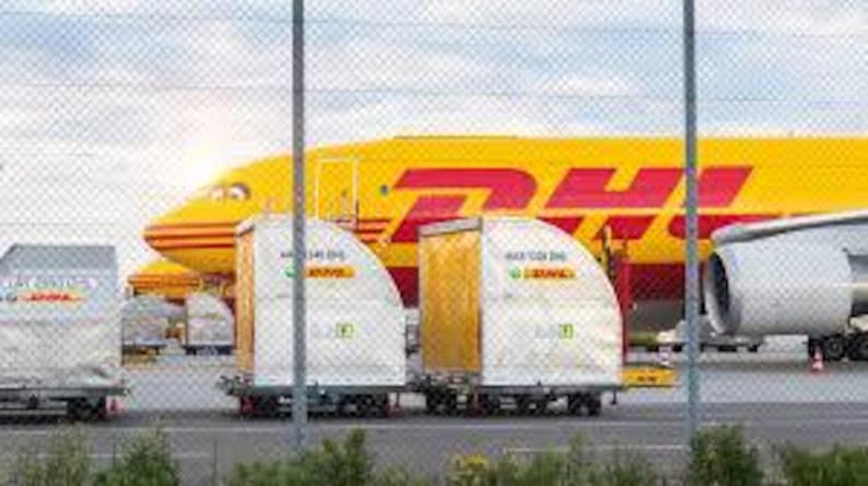 May include: A yellow DHL cargo plane with red lettering is parked on the tarmac. In front of the plane are several white and yellow cargo containers with the DHL logo. A chain-link fence is in the foreground.