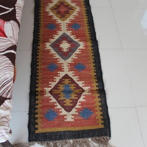 May include: A red, blue, and beige patterned rug with a black border and fringe. The rug has a geometric design with diamond shapes.