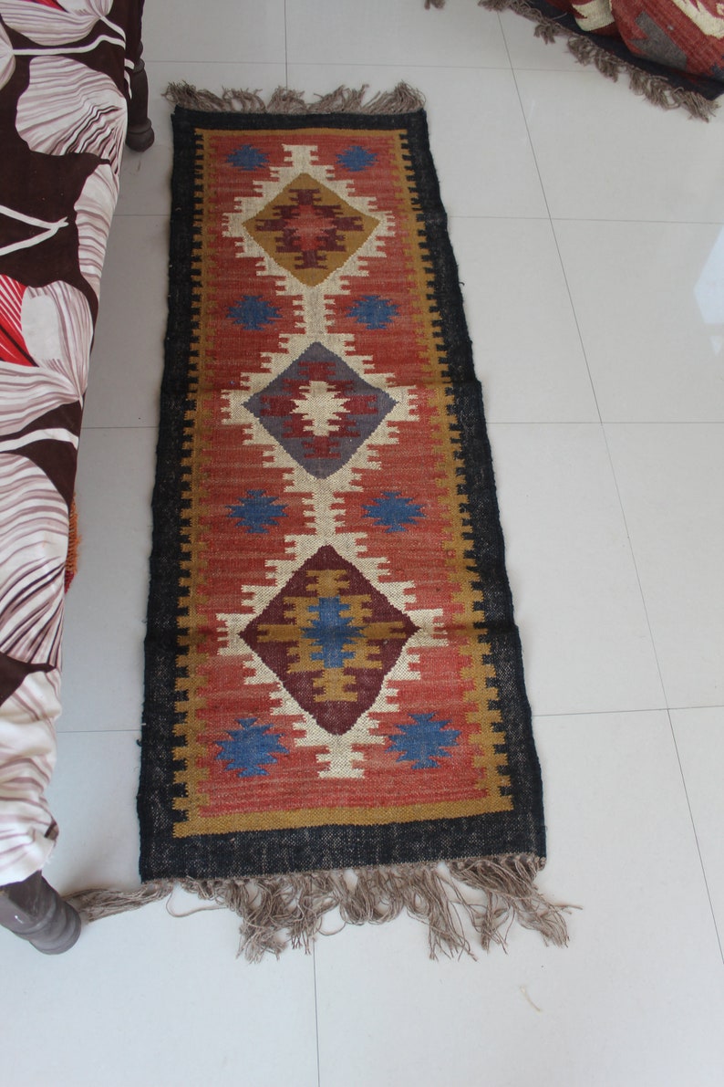 May include: A red, blue, and beige patterned rug with a black border and fringe. The rug has a geometric design with diamond shapes.