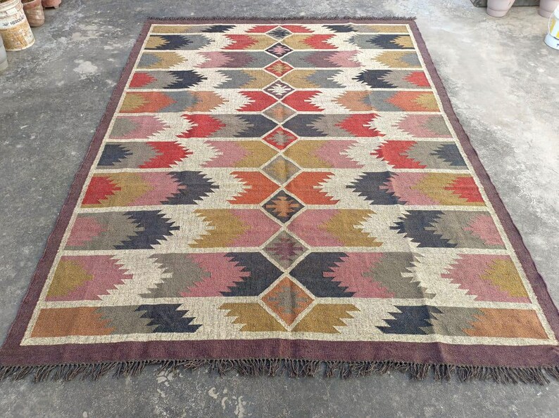 May include: A colorful geometric patterned rug with a brown border and fringe. The rug features a diamond pattern with shades of red, pink, orange, yellow, gray, and black.