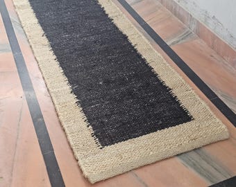 May include: A rectangular jute runner rug with a black center and a natural beige border. The rug is made of woven jute fibers, creating a textured surface. The runner is designed for hallways or entryways.
