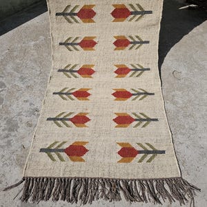 Könnte beinhalten: Ein beiger Jute-Teppich mit einem geometrischen Muster aus Pfeilen und Blumen in Rot-, Orange- und Grüntönen. Der Teppich hat einen Fransenrand.