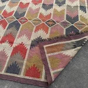 May include: A colorful geometric patterned rug with a fringe border. The rug is made of woven wool and features shades of red, pink, brown, and gray.