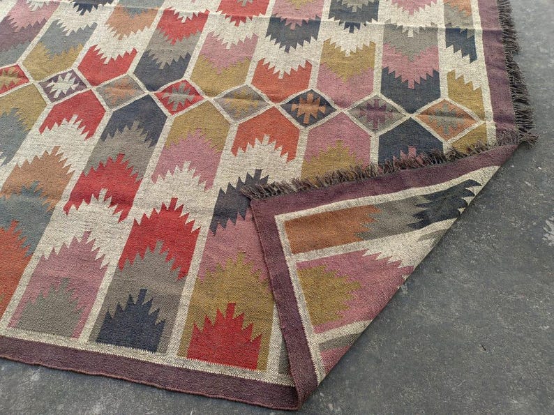 May include: A colorful geometric patterned rug with a fringe border. The rug is made of woven wool and features shades of red, pink, brown, and gray.