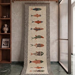 May include: A long, rectangular woven runner rug featuring a beige base with a repeating black geometric border and fringe at both ends. The centre displays a vertical column of ten stylised fish in various colours and patterns.