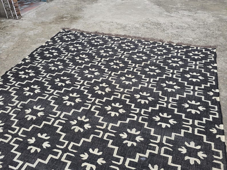 May include: A black and white patterned rug with a geometric design. The rug features a repeating pattern of white shapes on a black background. The rug has a fringed edge and is suitable for indoor use.