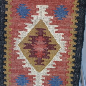 May include: A colorful rug with a geometric pattern in shades of red, blue, white, brown, and gold. The rug has a black border and fringe.