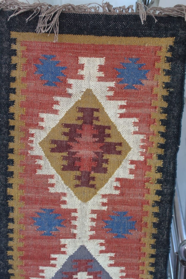 May include: A colorful rug with a geometric pattern in shades of red, blue, white, brown, and gold. The rug has a black border and fringe.