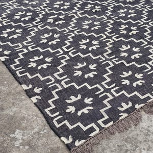 May include: Dark gray and white patterned rug with a geometric design. The rug features a repeating pattern of white shapes on a dark gray background. The rug has a fringed edge.