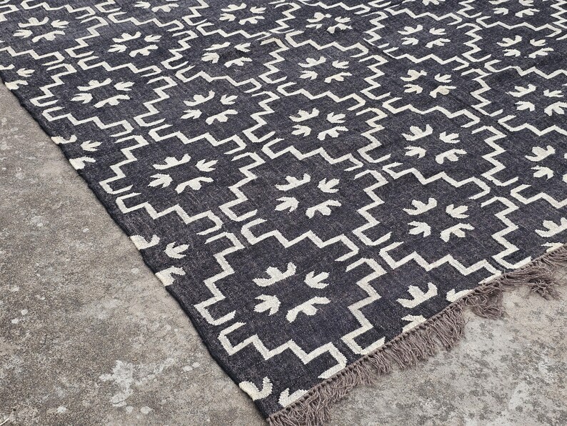 May include: Dark gray and white patterned rug with a geometric design. The rug features a repeating pattern of white shapes on a dark gray background. The rug has a fringed edge.