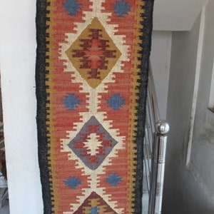 May include: A long, narrow rug with a black border and a colorful geometric pattern in shades of red, blue, brown, and white. The rug has a fringe on both ends.