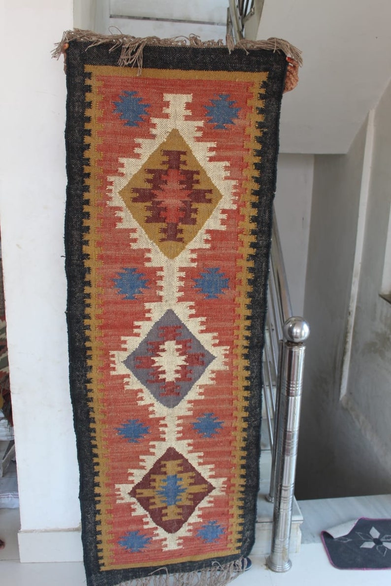 May include: A long, narrow rug with a black border and a colorful geometric pattern in shades of red, blue, brown, and white. The rug has a fringe on both ends.