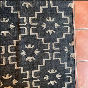 May include: A dark gray rug with a geometric pattern. The design features repeating light beige shapes, including stepped lines and stylized floral motifs. The rug's texture is visible, and it is placed next to a terracotta tile floor.