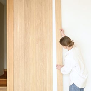Könnte beinhalten: Eine Person installiert hellfarbene Holzpaneele an einer Wand. Die Paneele sind vertikal und haben eine natürliche Holzmaserung. Die Person, bekleidet mit einem weißen Hemd und blauen Jeans, hält eine Platte an einer weißen Wand.