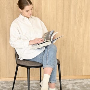 May include: A person sitting on a black metal chair with a gray fabric seat, reading a book. The person is wearing a white button-down shirt and blue jeans. The chair is in front of a wood panel wall.
