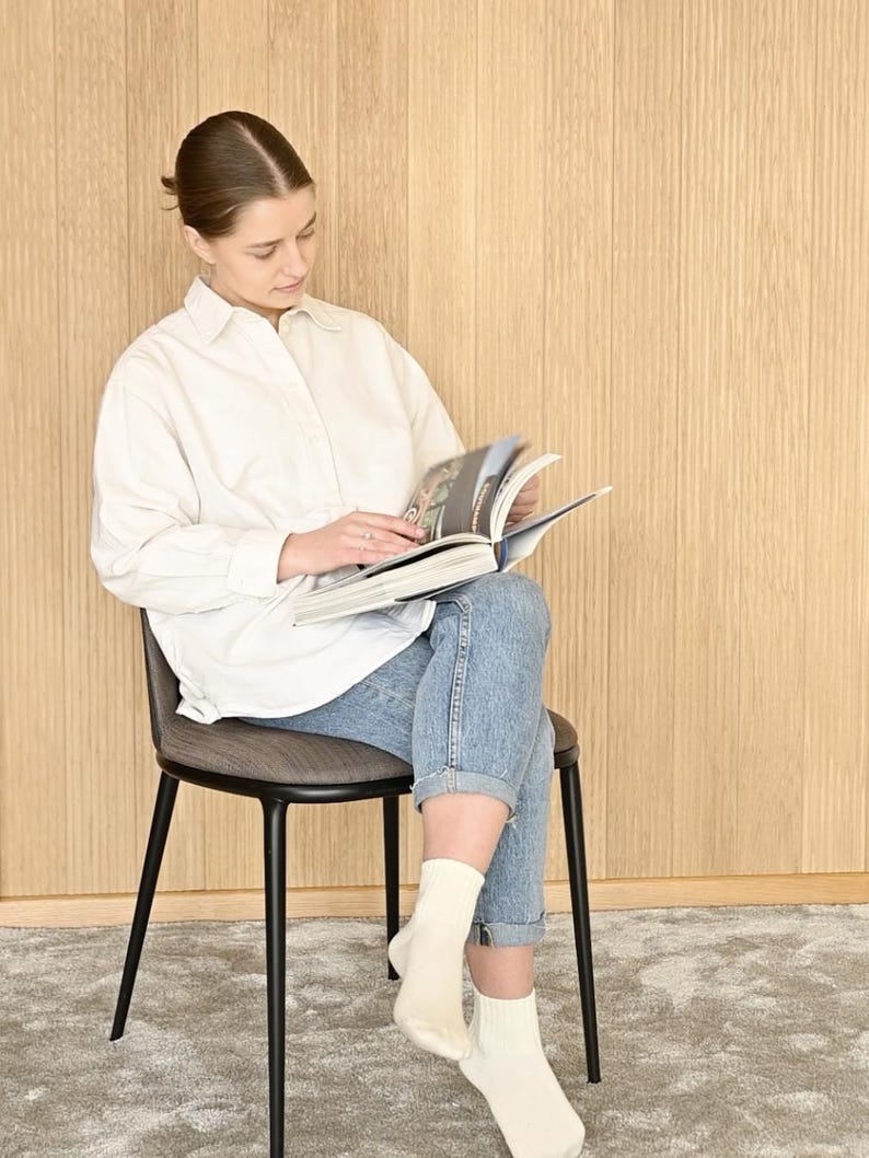 May include: A person sitting on a black metal chair with a gray fabric seat, reading a book. The person is wearing a white button-down shirt and blue jeans. The chair is in front of a wood panel wall.
