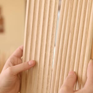 May include: Close-up of a light brown wood panel with vertical grooves. The panel is being held by two hands.