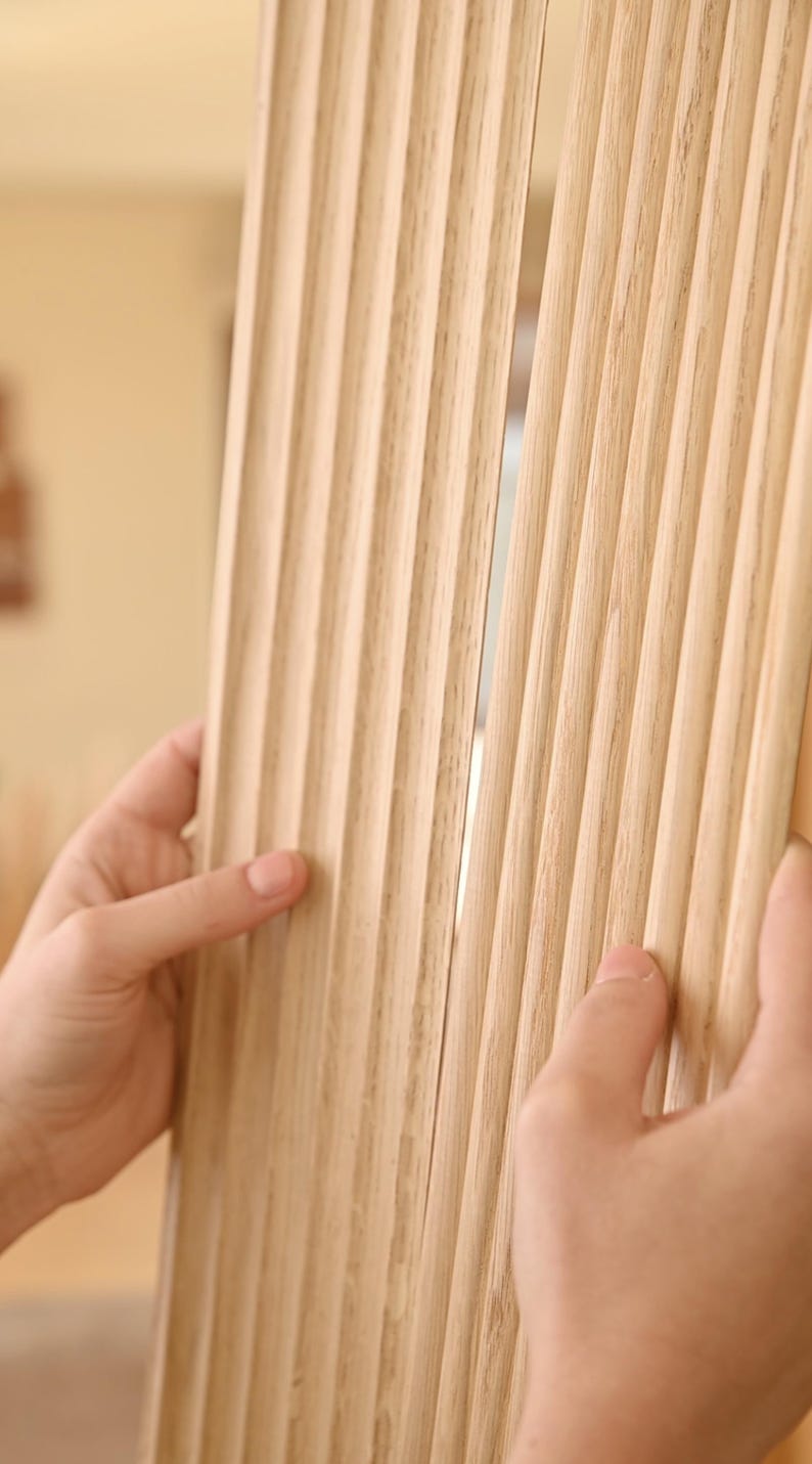 May include: Close-up of a light brown wood panel with vertical grooves. The panel is being held by two hands.