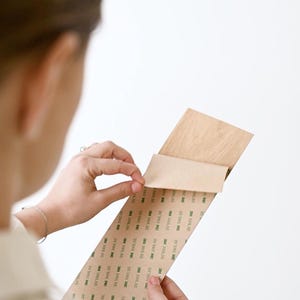 May include: A person is holding a wooden board with a brown adhesive tape on the back. The tape has the text "3M 300LSE" printed on it.