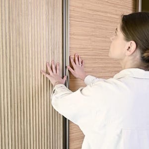 May include: A close-up of a wooden sliding door with a grooved texture. The door is partially open, revealing a similar wood panel on the other side. A person's hand is reaching out to touch the door.