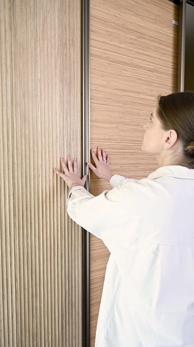 May include: A close-up of a wooden sliding door with a grooved texture. The door is partially open, revealing a similar wood panel on the other side. A person's hand is reaching out to touch the door.