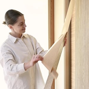 K&ouml;nnte beinhalten: Eine Person bringt eine selbstklebende Holzimitationfolie auf eine T&uuml;r an. Die Folie ist beige und hat eine Holzmaserung. Die T&uuml;r ist aus Holz und hat ein vertikales Paneeldesign.