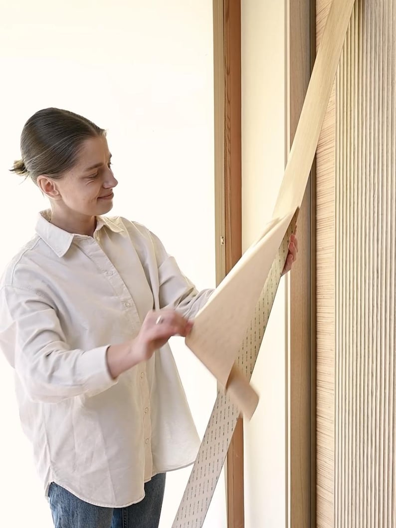K&ouml;nnte beinhalten: Eine Person bringt eine selbstklebende Holzimitationfolie auf eine T&uuml;r an. Die Folie ist beige und hat eine Holzmaserung. Die T&uuml;r ist aus Holz und hat ein vertikales Paneeldesign.