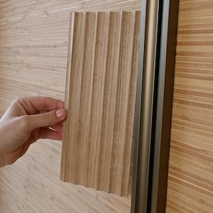 May include: A close-up of a hand holding a sample of wood paneling with a ribbed texture. The paneling is a light brown color and is being held up against a wall covered in a light brown woven bamboo wallpaper.