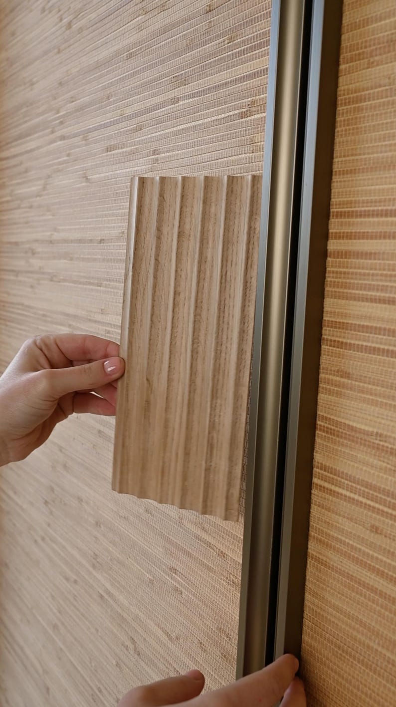 May include: A close-up of a hand holding a sample of wood paneling with a ribbed texture. The paneling is a light brown color and is being held up against a wall covered in a light brown woven bamboo wallpaper.