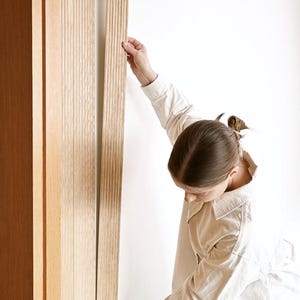 Könnte beinhalten: Eine Person montiert eine Holzleiste für eine Tür. Die Leiste ist aus hellem Holz und hat eine vertikale Maserung. Die Person trägt ein weißes Hemd und eine blaue Jeans.